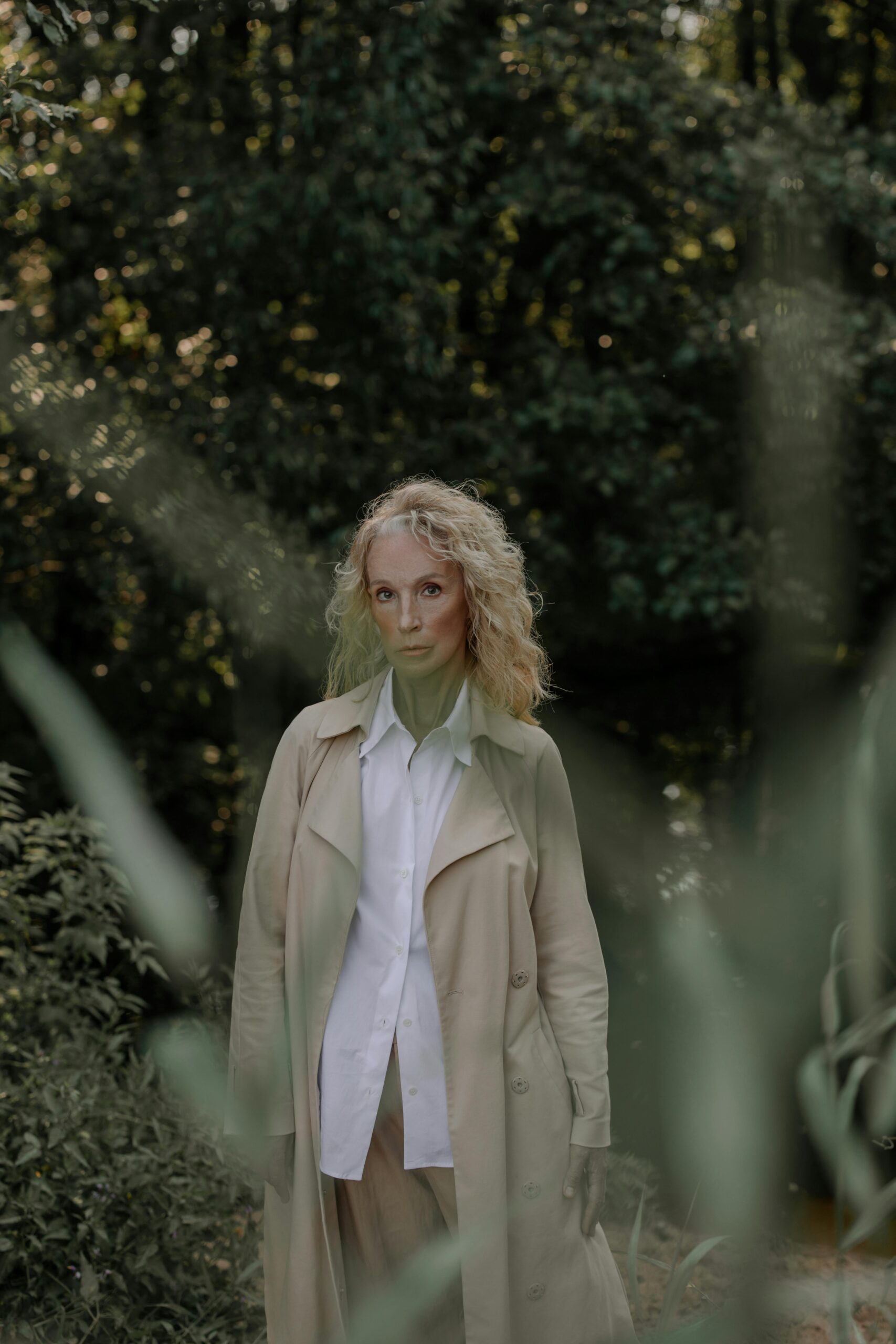 Blonde woman in a trench coat standing amidst lush greenery, exuding elegance.
